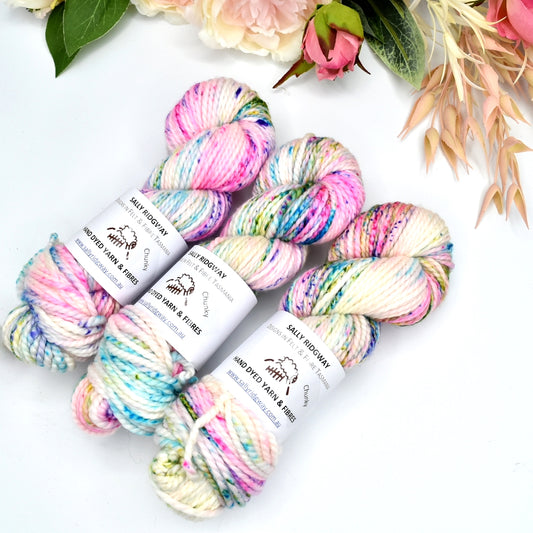 3 hanks of hand dyed speckled rainbow chunky knitting yarn laying side by side on a white background with flowers behind on an angle Buy hand dyed yarn online