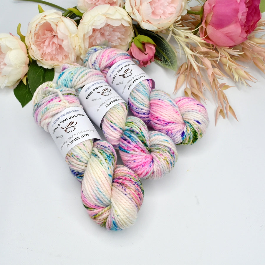 3 hanks of hand dyed speckled rainbow chunky knitting yarn laying side by side on a white background with flowers behind Shop online hand dyed yarn 