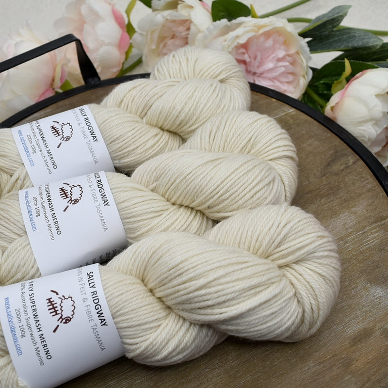Hand dyed knitting yarn in cream laying sideways on a timber tray with flowers. Shop 8 ply yarn online 